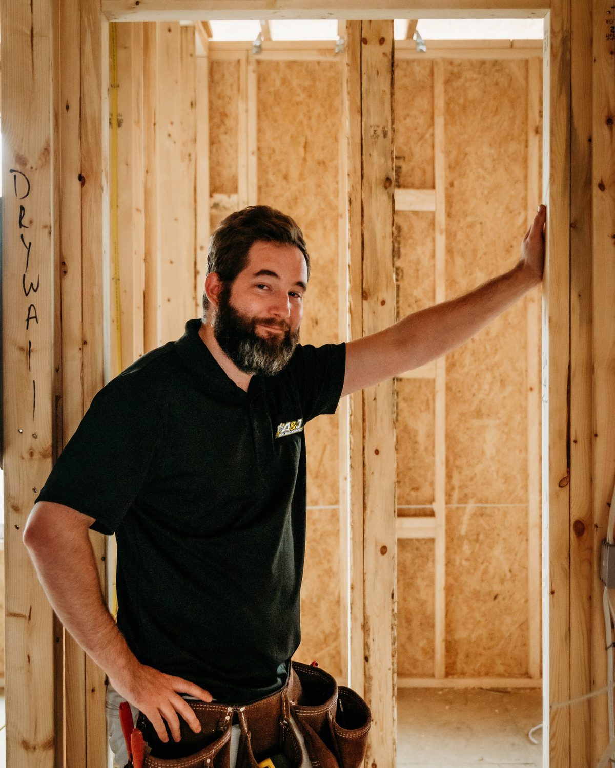 Adam Boyd leaning on a wall stud at a new-construction job, wearing an A&J Electric shirt and tool belt